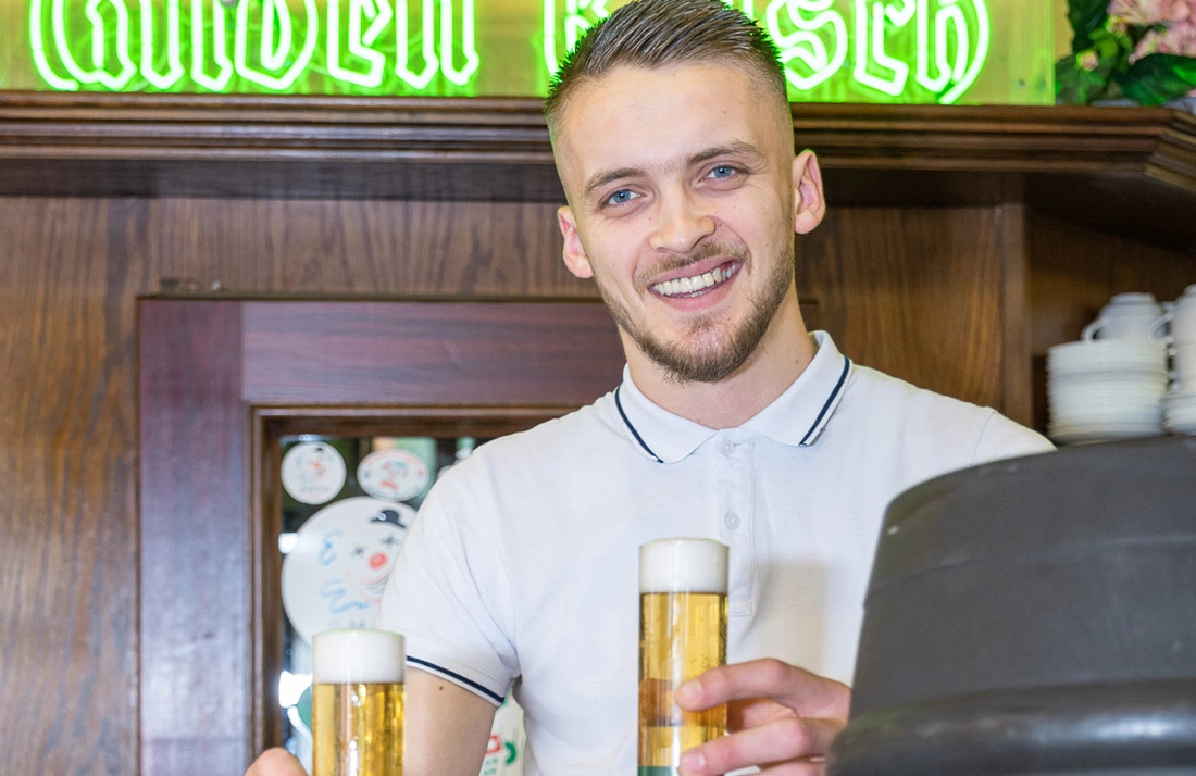 Ein lächelnder Kellner reicht zwei Gläser Bier über eine Theke.