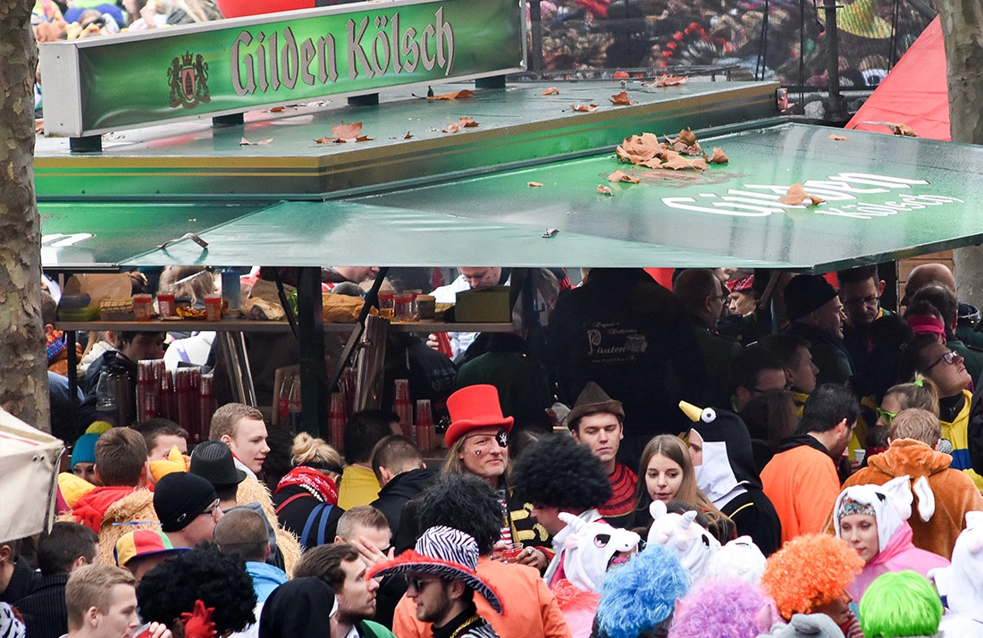 Menschen in farbenfrohen Kostümen versammeln sich um einen Getränkestand auf einem Fest.