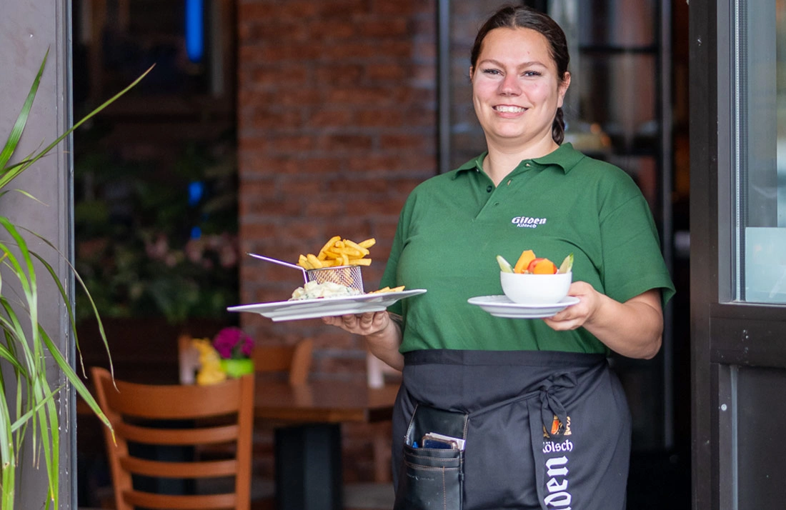 Eine Kellnerin in grüner Kleidung trägt zwei Teller mit Essen.