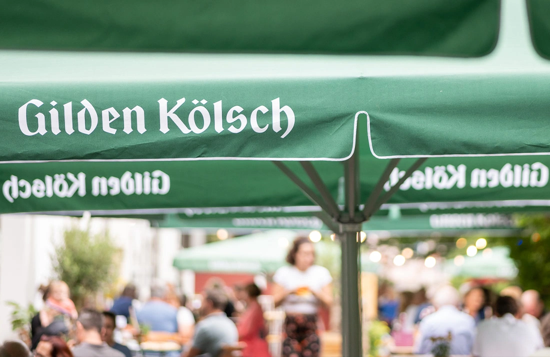 Grüner Sonnenschirm mit weißer Schrift über einem belebten Außenbereich eines Cafés.