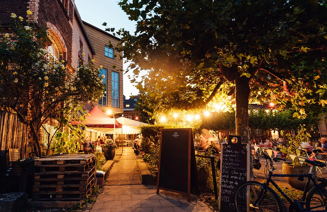 Ein belebter Außenbereich eines Restaurants bei Dämmerung mit Lichterketten in Bäumen und Gästen an Tischen.
