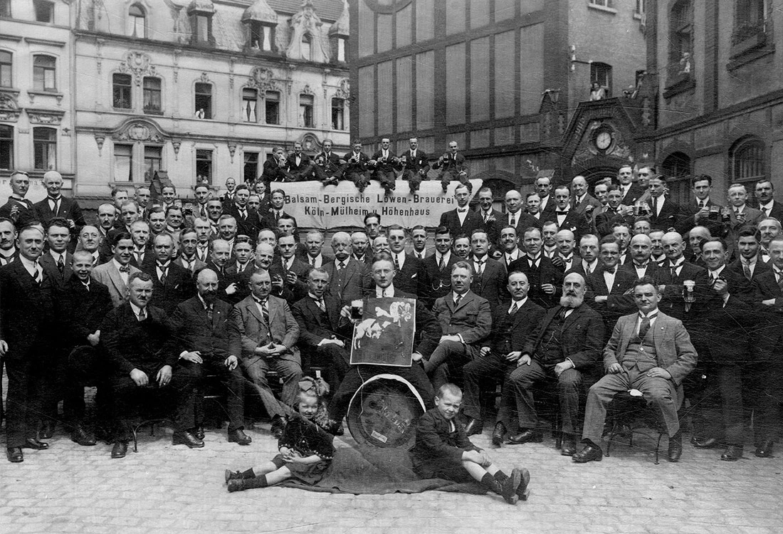 Eine Gruppe von über fünfzig Männern posiert vor einem historischen Gebäude, einige sitzen, die meisten stehen. Zwei Kinder sitzen im Vordergrund auf dem Boden.