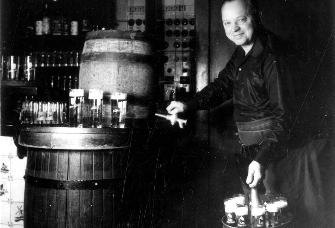 Ein Mann steht in einer Bar neben einem Holzfass und hält ein Tablett mit Gläsern.