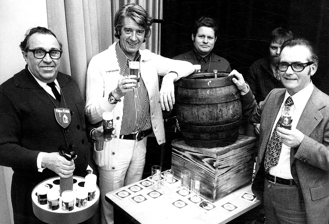 Vier Männer stehen um ein altes Holzfass herum und halten Biergläser. Einige tragen traditionelle Abzeichen.
