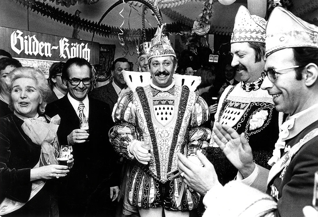 Fünf fröhliche Menschen in traditionellen Karnevalskostümen lachen und klatschen.