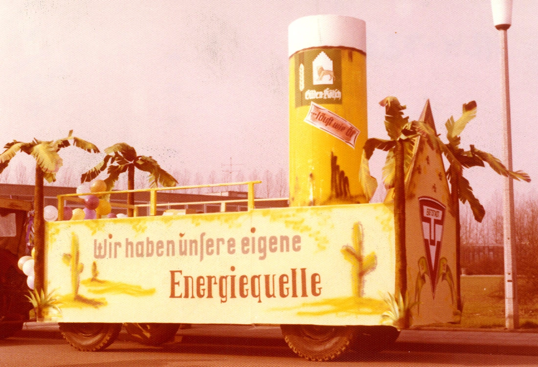 Ein Karnevalswagen mit großem, dekorativem Getränkebehälter und Palmen wird gezeigt. Auf einem Banner steht ein Slogan über Energiequellen.