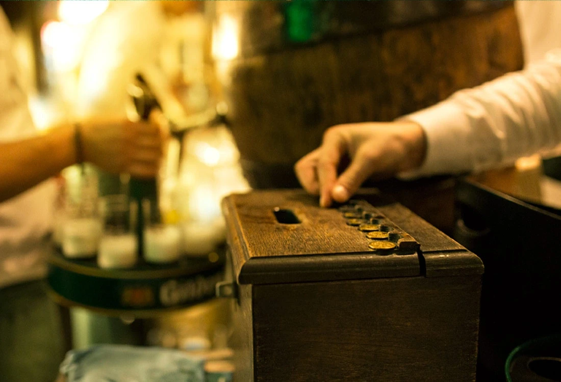Eine Person legt Münzen in Schlitze eines hölzernen Kastens an einer Bar.