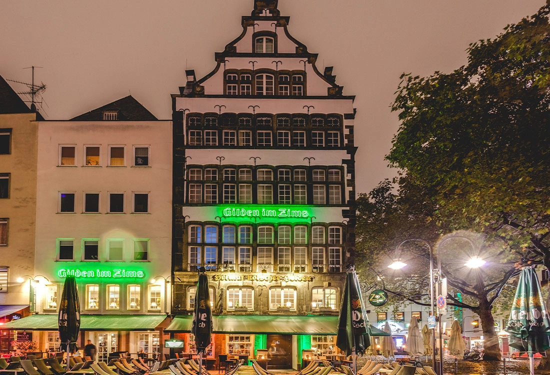 Historische und moderne Gebäude bei Nacht beleuchtet, darunter ein Café mit leeren Sitzgelegenheiten.
