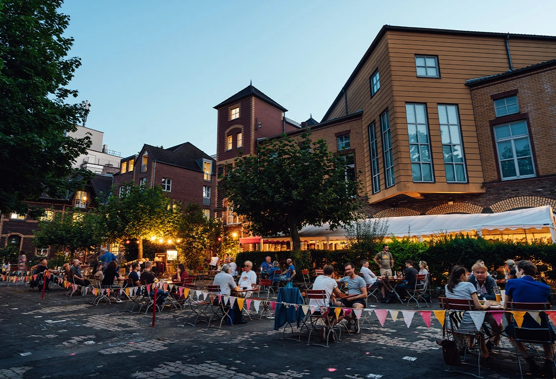 Menschen sitzen in einem belebten Freiluftcafé umgeben von alten Gebäuden am Abend.