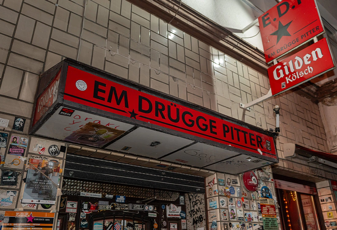 Ein rotes Leuchtschild mit Aufschrift über einem Eingang in einer gefliesten Wand, beklebt mit vielen Stickern.