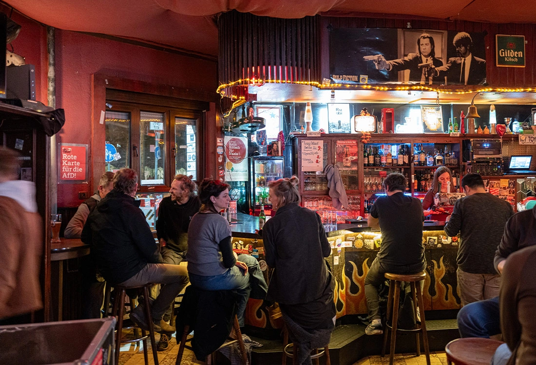 Mehrere Personen sitzen und unterhalten sich in einer belebten Bar. Die Bar ist warm beleuchtet und dekorativ eingerichtet.