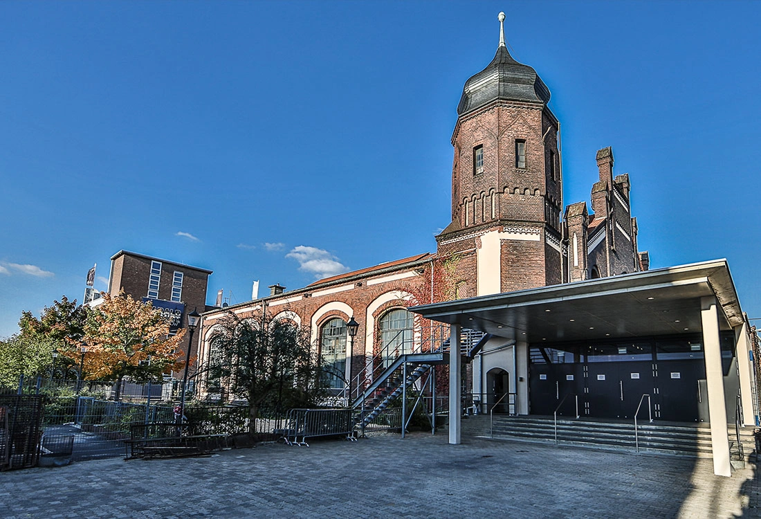 Ein historisches Gebäude mit einem großen Turm steht unter blauem Himmel. Nebenan ist ein moderner Anbau mit Überdachung.
